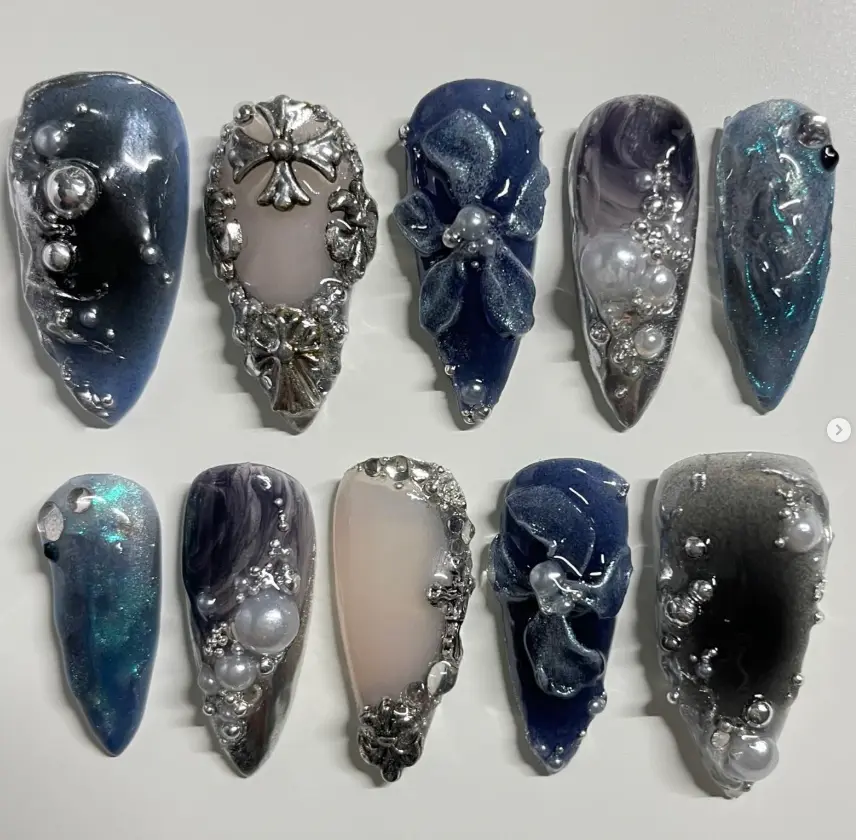 Elegant celestial nail art set in deep navy and silver tones, adorned with pearls, metallic crosses, sculpted flowers, and shimmering cosmic textures