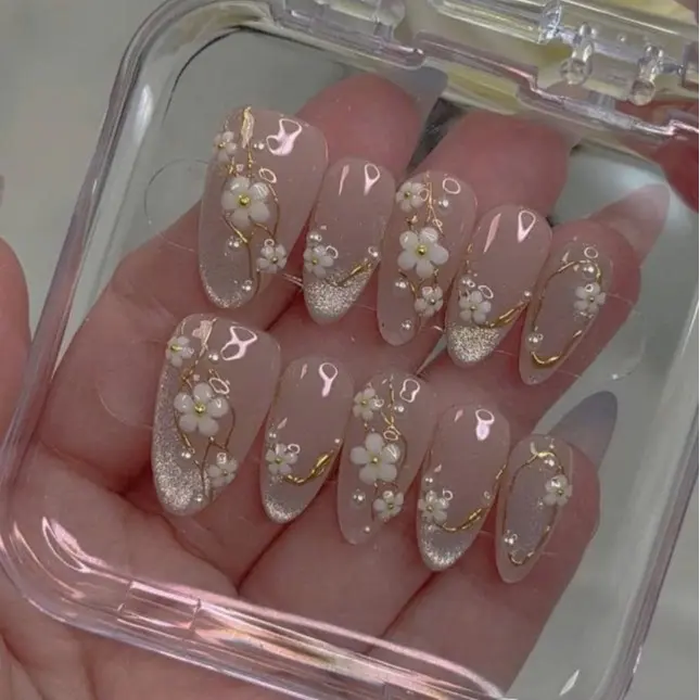 Luxurious nude almond press-on nails with glitter French tips and 3D white floral pearl accents, elegant bridal wedding nail art set with gold vine details for romantic brides